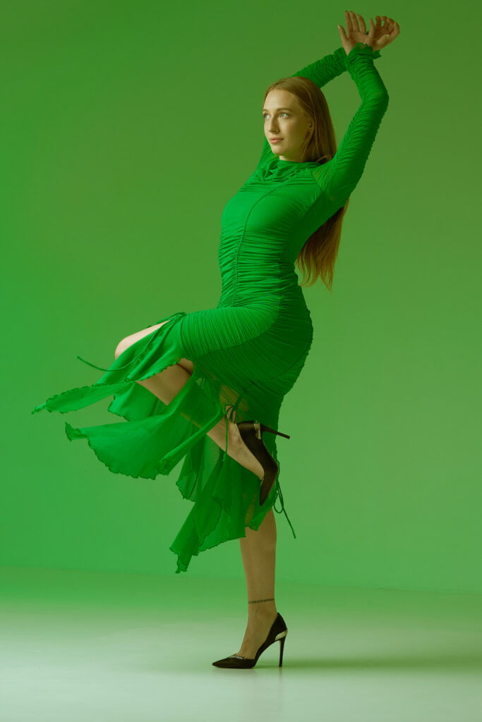 Photograph of a girl wearing a vibrant green dress, captured mid-jump against a matching green background. Her pose is dynamic and intriguing, showcasing her joyful energy as she leaps into the air. The green tones create a harmonious and captivating visual, emphasizing the girl's lively movement and the seamless connection between her attire and the environment.