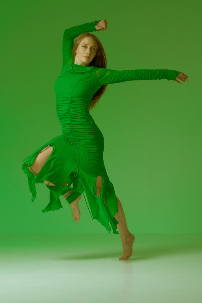 Photograph of a girl wearing a vibrant green dress, captured mid-jump against a matching green background. Her pose is dynamic and intriguing, showcasing her joyful energy as she leaps into the air. The green tones create a harmonious and captivating visual, emphasizing the girl's lively movement and the seamless connection between her attire and the environment.