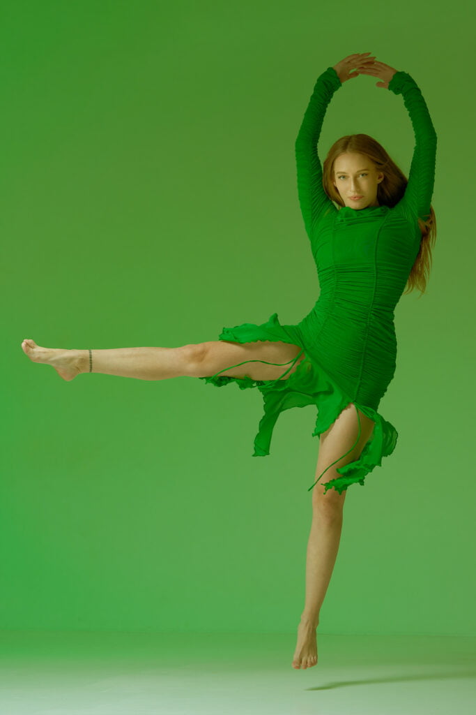 Photograph of a girl wearing a vibrant green dress, captured mid-jump against a matching green background. Her pose is dynamic and intriguing, showcasing her joyful energy as she leaps into the air. The green tones create a harmonious and captivating visual, emphasizing the girl's lively movement and the seamless connection between her attire and the environment.